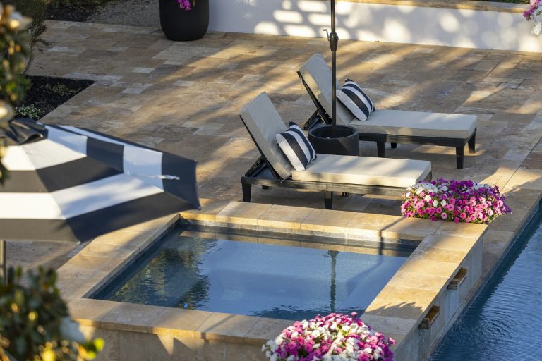Raised spa with travertine coping spilling into a backyard pool beside lounge seating