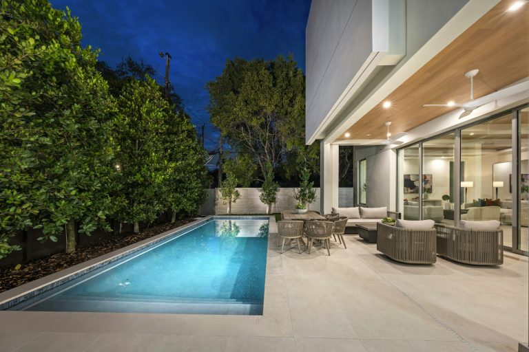 Narrow backyard pool bordered by a tall hedge beside a covered patio at dusk